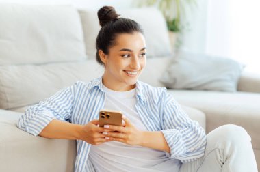 Kafkasyalı mutlu esmer kadın, evde dinleniyor, oturma odasındaki kanepenin yanında oturuyor, arkadaşlarıyla internet üzerinden akıllı telefon mesajları atıyor, online mağazalara bakıyor, e-postaları cevaplıyor, yüzünü çeviriyor, gülümsüyor.