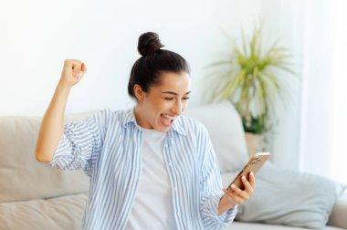 Neşeli, mutlu, beyaz bir kadın, elinde akıllı bir telefonla iyi haberler okuyor, zaferi kutluyor, maçı kazanıyor, kanepede oturuyor, yumruğunu sallıyor, gülümsüyor.