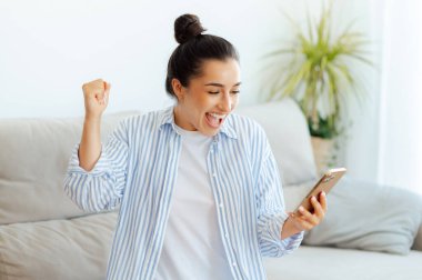 Heyecanlı genç Hintli ya da Arap kadın cep telefonuyla sosyal medyada sohbet ediyor, iyi haberler okuyor, zaferi kutluyor, evdeki kanepede oturup yumruklarını sallıyor, gülümsüyor.