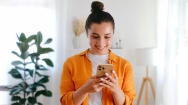Ofiste ya da evde dururken cep telefonu tutan turuncu tişörtlü mutlu Arap ya da Hintli genç kadın, sosyal medyada mesajlaşıyor, internette geziniyor, uygulamalarla ürünler sipariş ediyor.