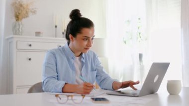 Mavi gömlekli mutlu Arap ya da Hintli iş kadını, proje yöneticisi, şirket CEO 'su, bir dizüstü bilgisayar üzerinde çalışıyor, strateji ve iş planı geliştiriyor, notlar alıyor, bilgi arıyor, gülümsüyor.