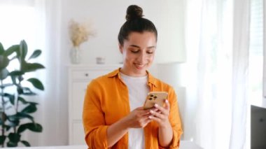 Cihazları kullanarak. Turuncu gömlekli, elinde akıllı telefon tutan, sosyal medyada mesaj yazan, internete göz gezdiren, e-postalarını kontrol eden pozitif Arap ya da Hintli genç bir kadının fotoğrafı.