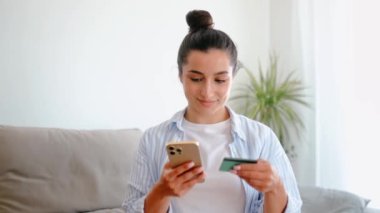 Pozitif çekici esmer bir kadın, oturma odasında bir koltukta oturuyor, akıllı telefon ve kredi kartı tutuyor, cep telefonu uygulamasından online ödeme için kart bilgilerini giriyor, online alışveriş yapıyor, gülümsüyor.