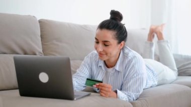 Güzel, mutlu beyaz kadın rahat bir kanepede dizüstü bilgisayarla oturuyor, elinde bir kredi kartı, mobil uygulama ile alışveriş yapıyor veya online bankacılık yapıyor, faturaları ödüyor, gülümsüyor.