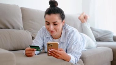 Gündelik giysiler içinde, bir kanepede uzanmış, akıllı telefon ve kredi kartıyla online alışveriş yapan internetten sipariş veren, faturaları ödeyen, gülümseyen beyaz bir kadın.