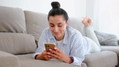 Mutlu genç Hintli ya da Arap güzel bir kadın evinde dinleniyor, kanepede uzanıyor ve cep telefonu kullanıyor arkadaşlarıyla mesajlaşıyor, online alışveriş yapıyor, haberlere çıkıyor, gülümsüyor.