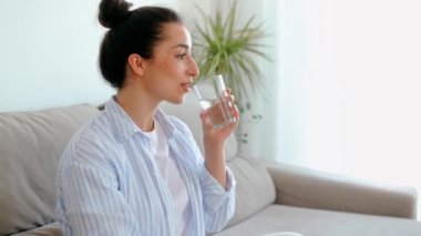 Mutlu pozitif Arap ya da Hintli esmer kadın sabahları oturma odasında bir kanepede otururken cam bardak temiz su içiyor. Sağlıklı yaşam tarzı kavramı