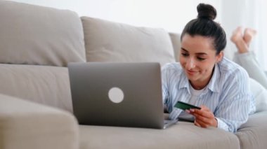 Çekici mutlu beyaz kadın gündelik giysiler içinde bir kanepede dizüstü bilgisayarla oturuyor, bir kredi kartı tutuyor, mobil uygulamalarla alışveriş yapıyor veya online bankacılık yapıyor, faturaları ödüyor, gülümsüyor.