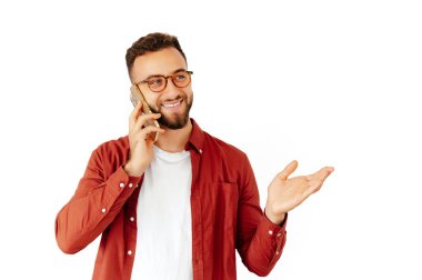 Gözlüklü, kırmızı tişörtlü, beyaz tişörtlü, neşeli, sakallı bir adam telefonda dostça gülümseyen ve el kol hareketleriyle konuşan beyaz bir arka planda izole edilmiş.