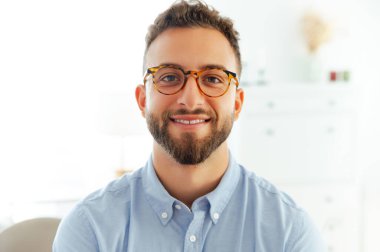 Gülümseyen, gözlüklü, mavi gömlekli, kendine güvenen ve profesyonel görünümlü başarılı bir adamın yakın plan portresi. Çekici, pozitif beyaz ya da Arap erkek ofiste kameraya bakıyor.