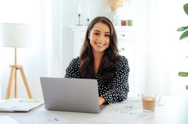 Kendine güveni tam, esmer bir kadın, ofis çalışanı, beyaz bir masada oturuyor, elinde belgelerle ve modern ofiste bir fincan kahveyle, kameraya bakıyor ve dostça gülümsüyor.