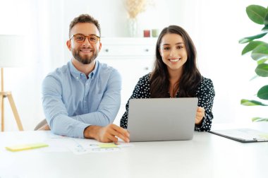 Pozitif çok ırklı iş arkadaşları modern bir ofiste çalışma masasında yan yana oturuyorlar. Önlerinde dizüstü bilgisayarlarıyla birlikte yeni bir proje üzerinde çalışıyorlar. Kameraya bakıyorlar, arkadaşça gülümsüyorlar.