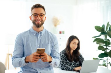 Mavi gömlekli ve gözlüklü yakışıklı bir iş adamı, elinde bir cep telefonu, arkadaşlarıyla ya da iş arkadaşlarıyla sohbet ediyor, ofiste duruyor ve kameraya gülümsüyor, arka planda dizüstü bilgisayarla çalışan bir kadın.