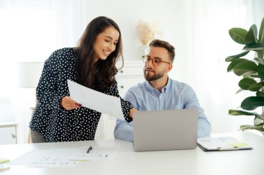 Profesyonel bir kadın bir rapor tutuyor ve gözlüklü bir adama sunuyor. Bir masada dizüstü bilgisayar ve veri kağıtlarıyla oturup gülümserken fikirleri tartışıyorlar.