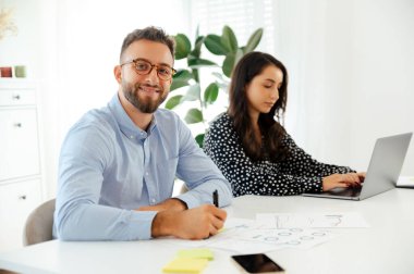 Mavi gömlekli, gözlüklü, bakışlı ve gülümseyen çekici iş adamı Arap ya da Kafkas kökenli biri ofiste oturuyor ve arka planda bir kadın dizüstü bilgisayarla dikkatlice çalışıyor.