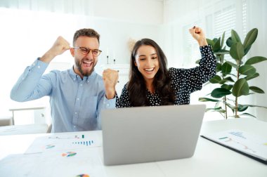 Masadaki başarıyı kutlayan neşeli iş ekibi, heyecan içinde yumruklarını kaldıran erkek ve kadın grafikleri analiz ediyor ve bir dizüstü bilgisayardaki hedeflere ulaşıyorlar, başarıyı ve işbirliğini simgeliyorlar.