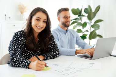 Mutlu, güzel, genç bir Arap ya da beyaz bir kadın masadaki yapışkan bir nota yazarken bir adam arka planda dizüstü bilgisayara yazarken kameraya gülümsüyor, takım çalışması ve yaratıcılık sergiliyor.