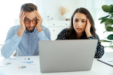 Stresli erkek ve kadın, modern bir çalışma alanında oturup, evrak ve çizelgelerle çevrili, endişe ve hayal kırıklığını gösteren kritik iş sonuçlarını inceleyen bir dizüstü bilgisayara bakmaktan şaşkına döndüler.