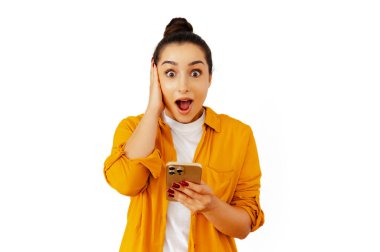 Sarı tişörtlü, elinde akıllı bir telefonla kameraya bakarken şaşkın ve şaşkın bir şekilde beyaz arka planda duran, beklenmedik haberlere şaşırmış ve şaşırmış genç esmer kadın.