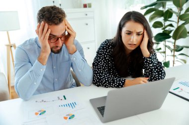 Stresli meslektaşlar bir dizüstü bilgisayardaki veri ve çizelgeleri analiz ediyor ve bir masada kağıt basıyor, modern bir ofiste otururken bir sorunu çözmek için birlikte çalışıyor, kafa karışıklığı ve hayal kırıklığı yaşıyor