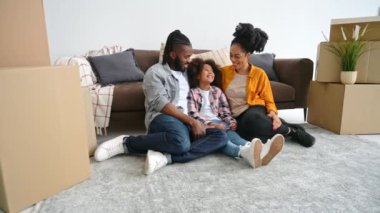 Yeni ev, taşınma. Mutlu Afro-Amerikan ebeveynler ve şirin küçük kızları, ellerinin sembolik çatısını yapıyorlar, birlikte oturma odasında koltuğun yanında oturuyorlar, kameraya bakıp gülümsüyorlar.