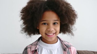 Güzel, Afro-Amerikan bir kızın video portresi, kreş çağında, kıvırcık saçlı, basit giyimli, kanepede oturuyor, kameraya bakıyor, arkadaşça gülümsüyor. Mutlu çocuk