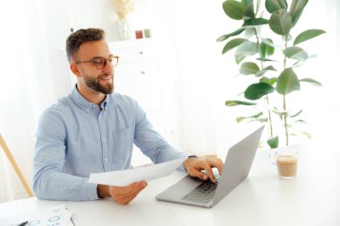 Kendine güveni tam bir iş adamı, elinde kağıtları parlak ve modern bir ofiste tutarken, bir projede başarı planları ve iş çizelgelerini gözden geçirirken, gülümserken bilgisayarında çalışır.