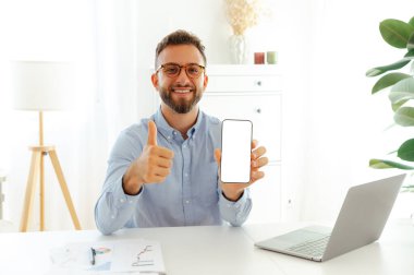 Mutlu iş adamı, sunumunuz ya da reklamınız için beyaz ekranlı bir akıllı telefonu kaldırıyor ve heyecan ve pozitiflik gösteren parlak, modern bir ofiste başparmak kaldırıyor.