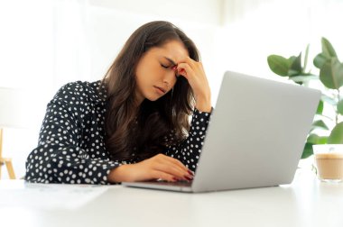 Sinirli iş kadını elinde dizüstü bilgisayarla masada oturuyor, yorgun ve çok çalışıyor görünüyor, masaya dağılmış kağıtlar ve yanında bir bardak kahveyle, baş ağrısı, kapalı gözler, dinlenmeye ihtiyacı var.