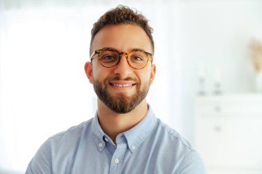 Gözlüklü başarılı bir beyaz ya da Arap işadamının yakın plan fotoğrafı, modern ofiste kendine güvenen, pozitifliğini ve yeni zorluklara hazır olduğunu gösteren, kameraya gülümseyen