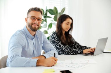 Gözlüklü bir işadamı, modern bir ofiste meslektaşı ile raporlar üzerinde çalışıyor, çizelgeler ve dizüstü bilgisayarla çevrili bir masada oturuyor, takım çalışması ve üretkenlik sergiliyor, kameraya gülümsüyor.