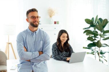 Çekici, kendine güvenen, gülümseyen Arap ya da beyaz, gözlüklü, mavi gömlekli, kolları çapraz duran, modern bir ofiste dizüstü bilgisayar üzerinde çalışan bir bayan.