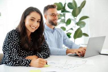 Mutlu, çekici, genç bir Arap ya da beyaz bir kadın masadaki yapışkan bir nota yazı yazıp kameraya gülümserken bir adam arka planda dizüstü bilgisayara yazarak takım çalışması ve yaratıcılık sergiliyor.