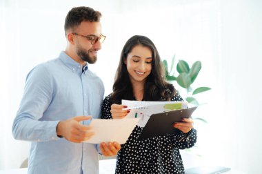 Mavi gömlekli ve gözlüklü başarılı bir adam ve esmer güzel bir kadın modern bir ofiste yan yana durup finansal belgeleri tutuyor ve bilgileri analiz ediyor, fikirleri tartışıyor, gülümsüyor.