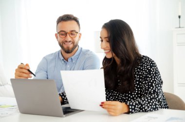 Modern ofisteki bir masada belgelerle çalışan iki başarılı iş arkadaşı. İkisi de gülümsüyor, dizüstü bilgisayarları ve çizelgeleri masada. Fikirlerini paylaşıyorlar, iş planı geliştiriyorlar.