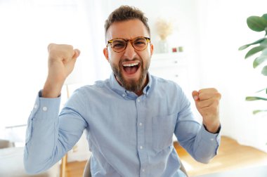Mavi gömlekli neşeli beyaz ya da Arap bir adam, heyecan içinde yumruklarını kaldırıyor, parlak, modern bir ofiste başarıyı kutluyor. Mutluluk, zafer ve kişisel başarı kavramı