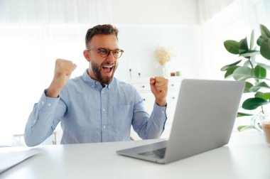 Mavi gömlekli neşeli başarılı iş adamı, dizüstü bilgisayarına bakarken yumruklarını kaldırıyor, parlak, modern bir ofiste başarısını kutluyor, mutlu bir şekilde gülümsüyor. Başarı hedefi