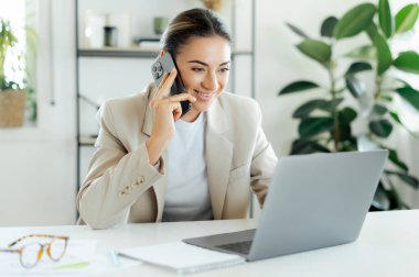 Cep telefonu sohbeti. Şirin beyaz iş kadını, şirket yöneticisi iş arkadaşı ya da müşterisiyle akıllı bir telefonla konuşuyor ve modern bir ofiste dizüstü bilgisayarla masasına notlar yazıyor.