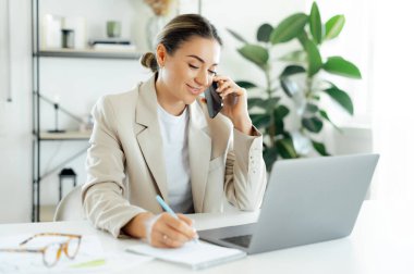 Gülümseyen Kafkas iş kadını, şirket yöneticisi cep telefonuyla konuşuyor ve masasına dizüstü bilgisayarla parlak modern bir ofiste notlar yazıyor, müşterisiyle akıllı telefon konuşması yapıyor.