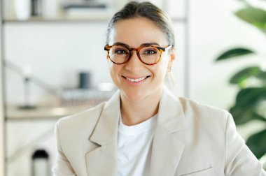 Modern ofis ortamında memnuniyet ve başarı gösteren gözlüklü mutlu beyaz iş kadınının yakın plan fotoğrafı. Kameraya bakıyor ve dostça gülümsüyor.