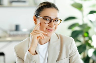 Modern ofis ortamında memnuniyet ve başarı gösteren gözlüklü, kendine güvenen beyaz bir iş kadınının yakın plan fotoğrafı. Kameraya bakıyor ve dostça gülümsüyor.