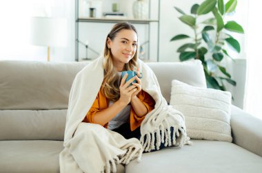 Sarı gömlekli, rahat bir battaniyeye sarılı, oturma odasındaki kanepede otururken elinde sıcak bir fincan çay tutan beyaz bir kadın. Rahatlama, rahatlama ve ev sağlığı kavramı