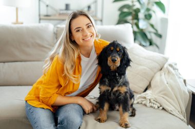 Günlük kıyafetler giymiş güzel beyaz bir kadın ve oturma odasında kanepede oturan sevimli siyah köpeği, ikisi de kameraya bakıyor, kadın arkadaşça gülümsüyor.