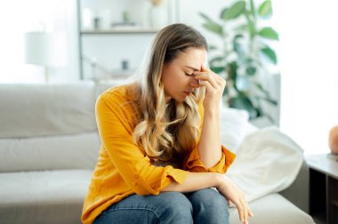 Stres, endişe, yorgunluk ve zihinsel yorgunluk kavramı. Sarı gömlekli, yorgun, beyaz bir kadın rahat bir koltukta oturuyor, burun köprüsüne masaj yapıyor, baş ağrısı ya da kişisel sorunları var.