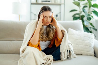 Baş ağrısı, stres ve yorgunluk kavramı. Sarı gömlekli bitkin, beyaz bir kadın oturma odasındaki bir kanepede battaniyeye sarılmış, şakaklarına masaj yapıyor, migreni tutuyor.