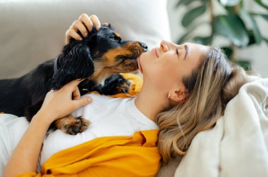 Neşeli beyaz kadın ve onun sevimli siyah köpeği oturma odasındaki kanepede sevgi dolu bir anı paylaşıyorlar. Köpek, gözleri kapalı gülümserken sevgiyle çenesini yalıyor ve bağlarının tadını çıkarıyor.