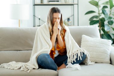 Sıcak bir battaniyeye sarılmış, mendile hapşıran, uzun sarı saçlı, hasta, beyaz bir kadın. Soğuk algınlığı, grip, mevsimsel hastalık ve alerji kavramı