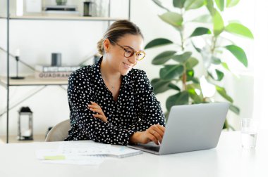 Gözlüklü, özgüvenli beyaz iş kadını, çalışma masasında dizüstü bilgisayarla oturuyor, klavyeye oturuyor, gülümsüyor ve modern bir ofiste finansal bir rapor üzerinde çalışıyor.