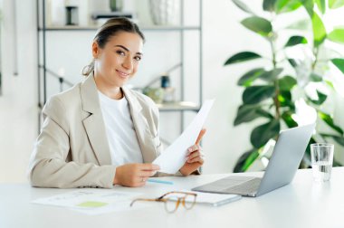 Beyaz takım elbiseli mutlu beyaz iş kadını elinde bir belge, elinde bir dizüstü bilgisayar ve modern ofis alanında bir bardak su olan bir masada oturuyor ve kameraya gülümsüyor.