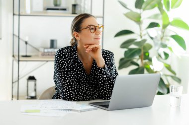 Düşünceli, kendine güvenen, gözlüklü beyaz bir iş kadını, dizüstü bilgisayarla bir masada oturuyor, başka yere bakıyor, düşünüyor ya da hayal kuruyor, modern bir ofiste çalışırken mola veriyor, gülümsüyor.